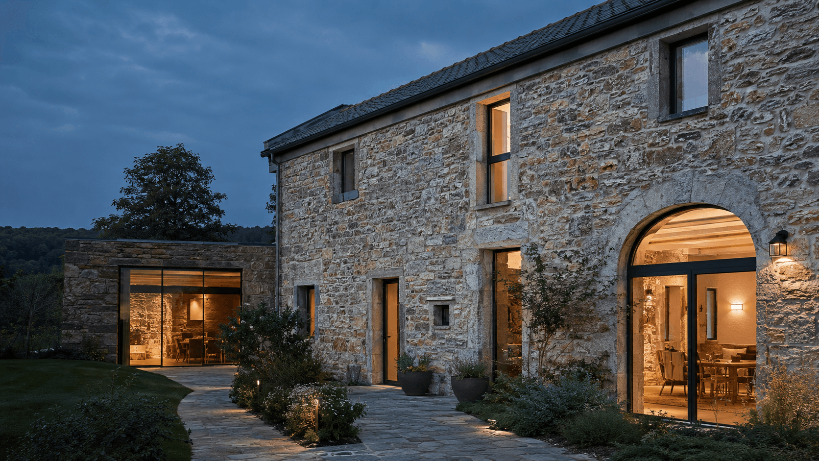 Longère en pierre rénovée avec extension bois et arche d'entrée éclairée, vue de nuit